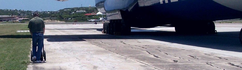 A surveyor pushing a ground-penetrating radar cart across an airport apron beside a parked aircraft in Antigua.