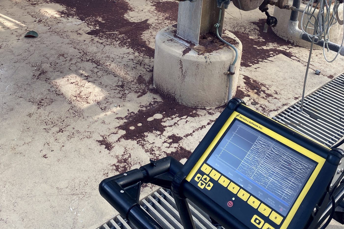 A ground-penetrating radar cart in a chemical plant, its screen showing a live radargram beside pipework and a concrete equipment pier.