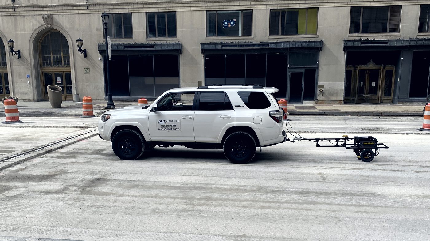 A GeoSearches vehicle towing a ground-penetrating radar cart along a closed downtown street during a utility survey.