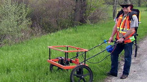 A surveyor in high-visibility clothing towing a low-frequency ground-penetrating radar antenna along a rural roadside.